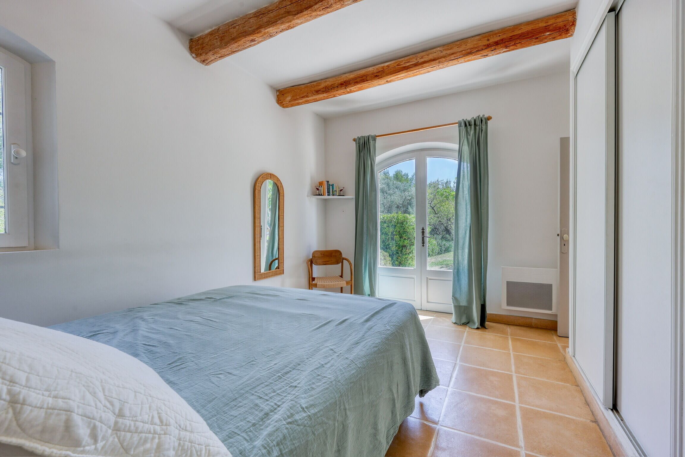 A bedroom with a bed, a wooden ceiling beam, a window with curtains, and a door leading to a balcony.