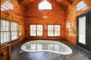 Interior de una cabaña de troncos con un jacuzzi, paredes de madera y ventanas amplias.