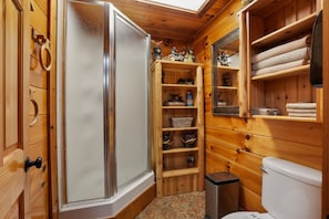 A compact wooden bathroom with a glass shower enclosure, wooden shelves with toiletries, and a white toilet.