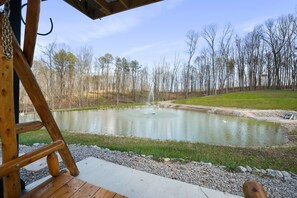 A wooden deck overlooking a pond with a fountain, surrounded by a grassy area and trees.