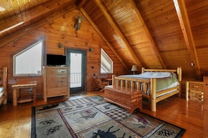 A cozy attic bedroom with wooden walls and ceiling, a bed, a dresser, a desk, and a large window.