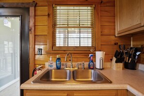A kitchen with wooden walls, a window with blinds, two sinks, and various kitchen items including a soap dispenser, a dish soap bottle, and a knife block.