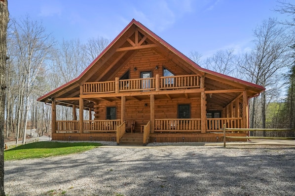 A wooden cabin with a balcony, a porch, and a gravel driveway.