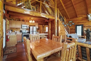 A wooden cabin interior with a dining area, kitchen, and a staircase leading to a loft.