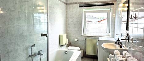 Un baño con ducha envuelta en vidrio, bañera, inodoro, lavamanos y ventana con vistas al exterior.