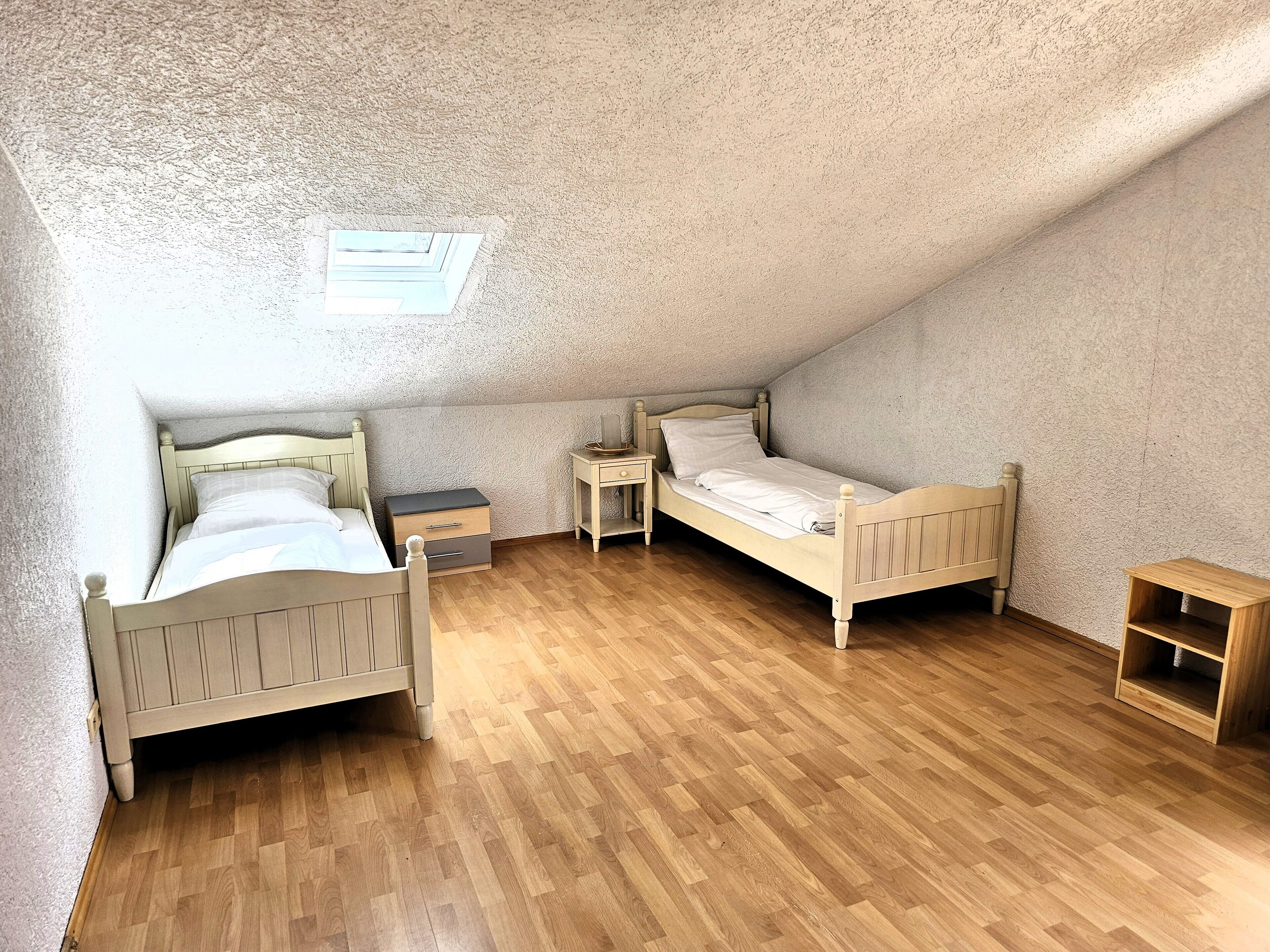 Un dormitorio pequeño con techo inclinado, dos camas de madera, una mesita de noche, un estante de madera pequeño y una pared con textura.