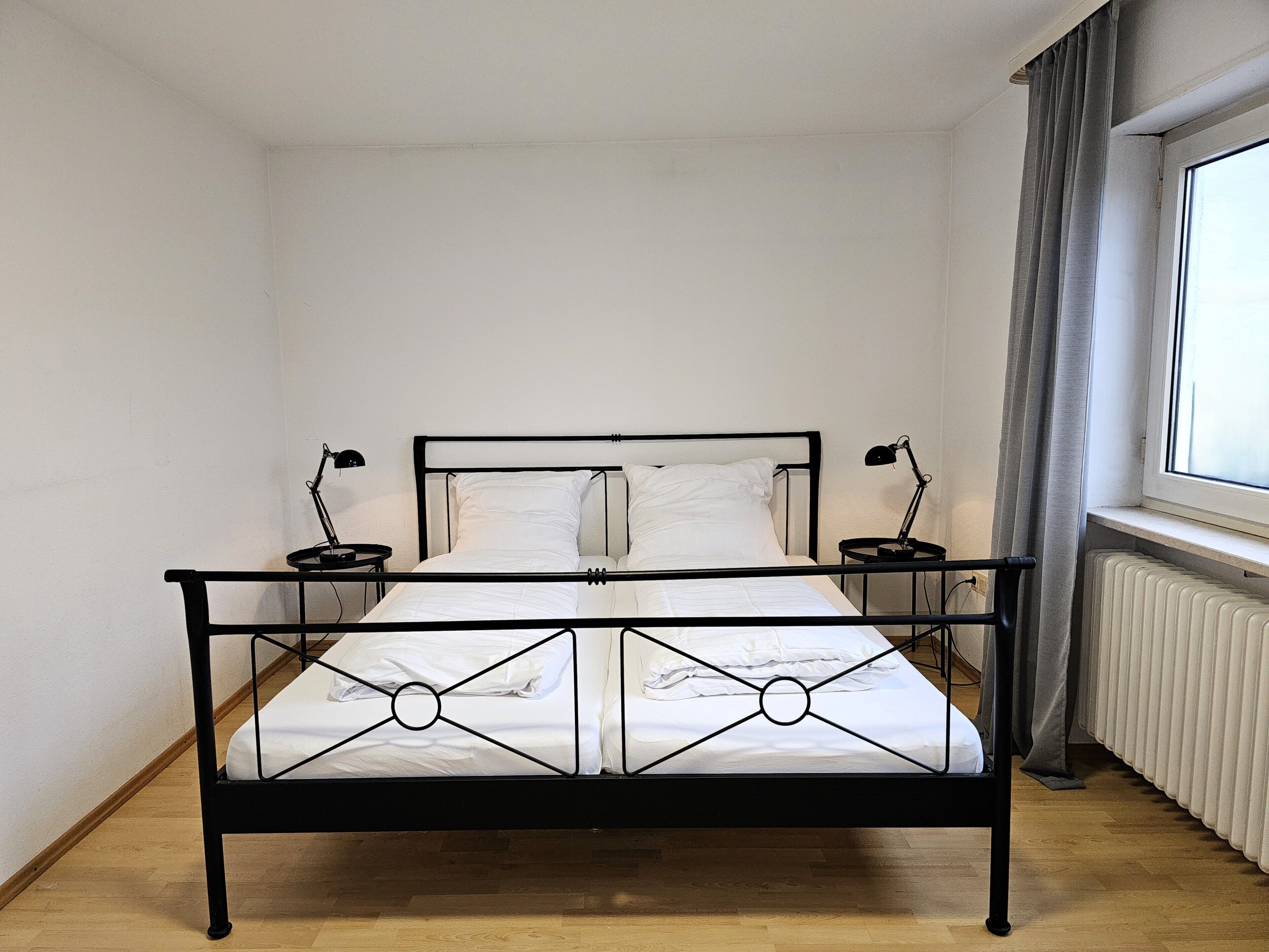 A single bed with a metal frame, two bedside tables, a window with curtains, and a radiator.