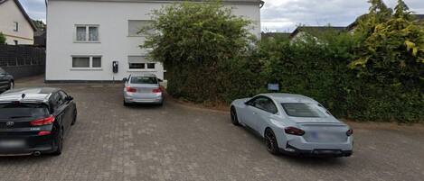 Uma entrada de garagem residencial com dois carros estacionados, uma casa branca com antena parabólica e um arbusto.