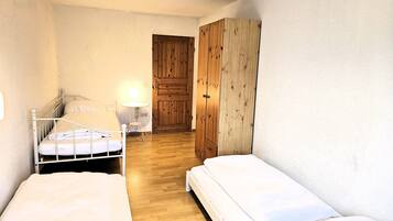 A small, single-room accommodation with two beds, a wooden wardrobe, a small table with a lamp, and a door leading to another room.