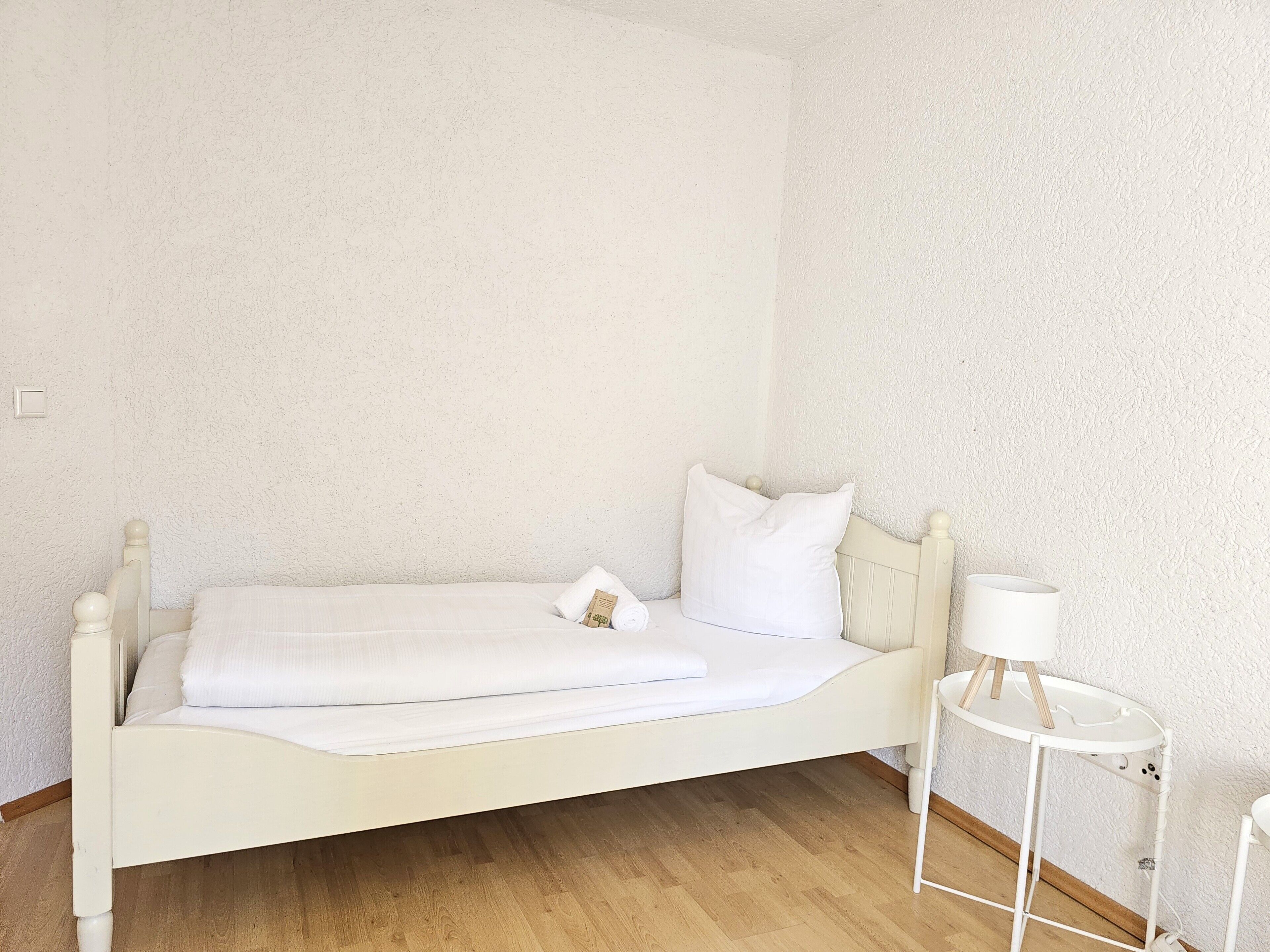 A single white bed with a wooden headboard and footboard, a small bedside table with a lamp, and a white wall.
