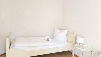 A single white bed with a wooden headboard and footboard, a small bedside table with a lamp, and a white wall.