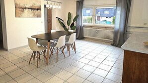 A dining area with a table and chairs, a potted plant, a radiator, and a window with curtains. - Apartmenthaus - Mackenbach (Mackenbach)
