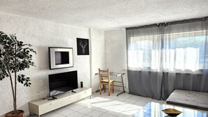 A modern living room with a flat-screen TV, a glass coffee table, a sofa, and a dining area with a chair and table. - Apartmenthaus - Mackenbach (Mackenbach)
