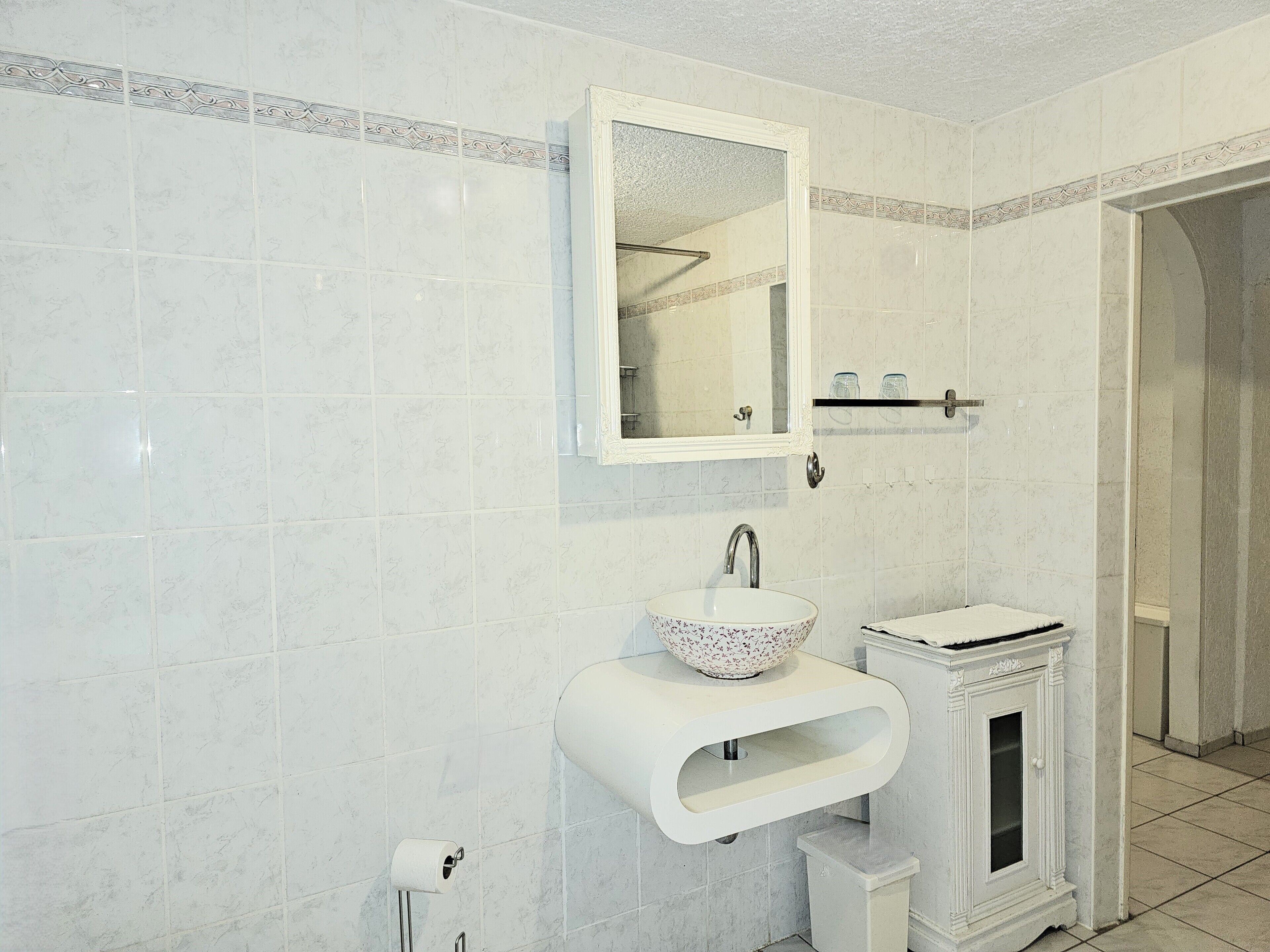 A bathroom with white tiles, a sink with a decorative bowl, a mirror, and a cabinet.