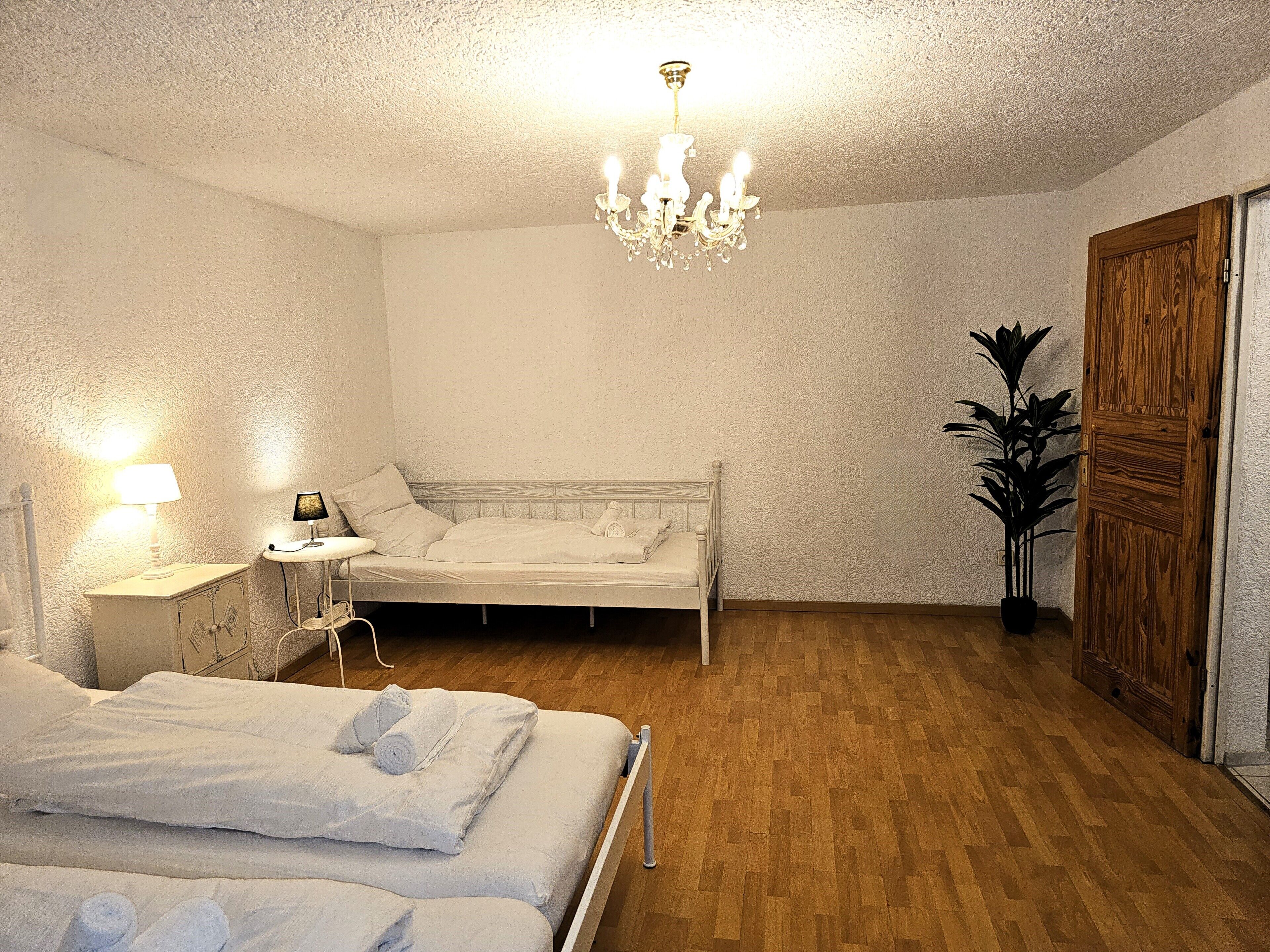 A bedroom with a wooden floor, a white bed, a bedside table with a lamp, and a chandelier.