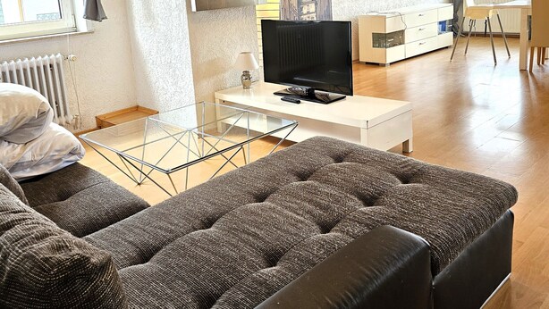 A modern living room with a large sectional sofa, a TV on a stand, a chandelier, and a dining area with a table and chairs. - Apartmenthaus - Mackenbach (Mackenbach)
