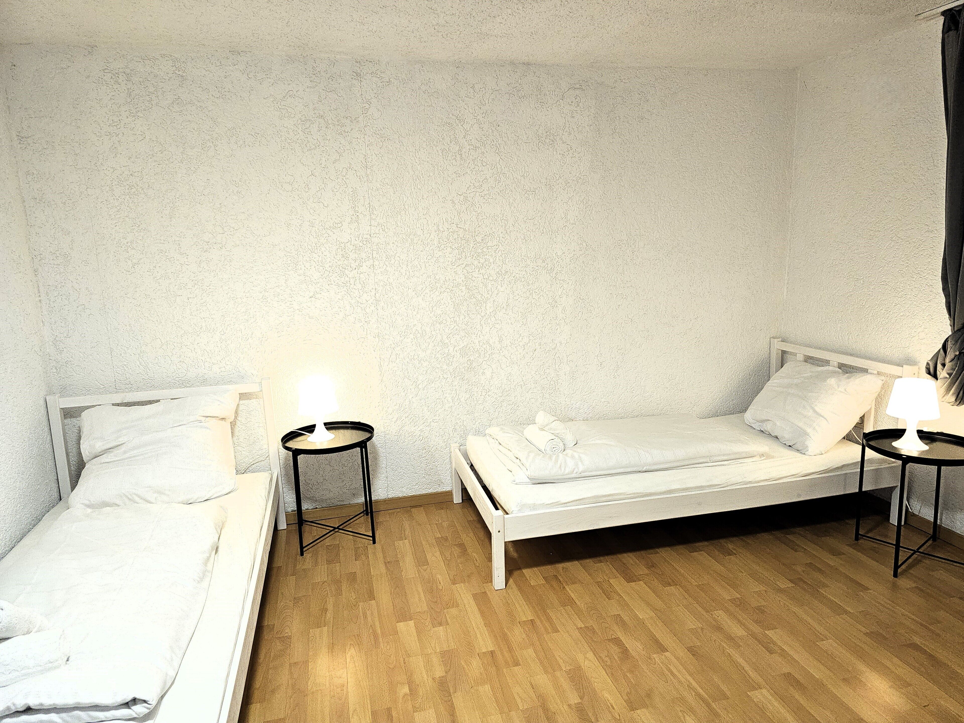 Il s’agit d’un petit logement composé d’une seule pièce, comprenant deux lits simples, chacun avec un oreiller et une petite table de chevet dotée d’une lampe. La pièce est agrémentée d’un mur à texture et d’un plancher en bois.