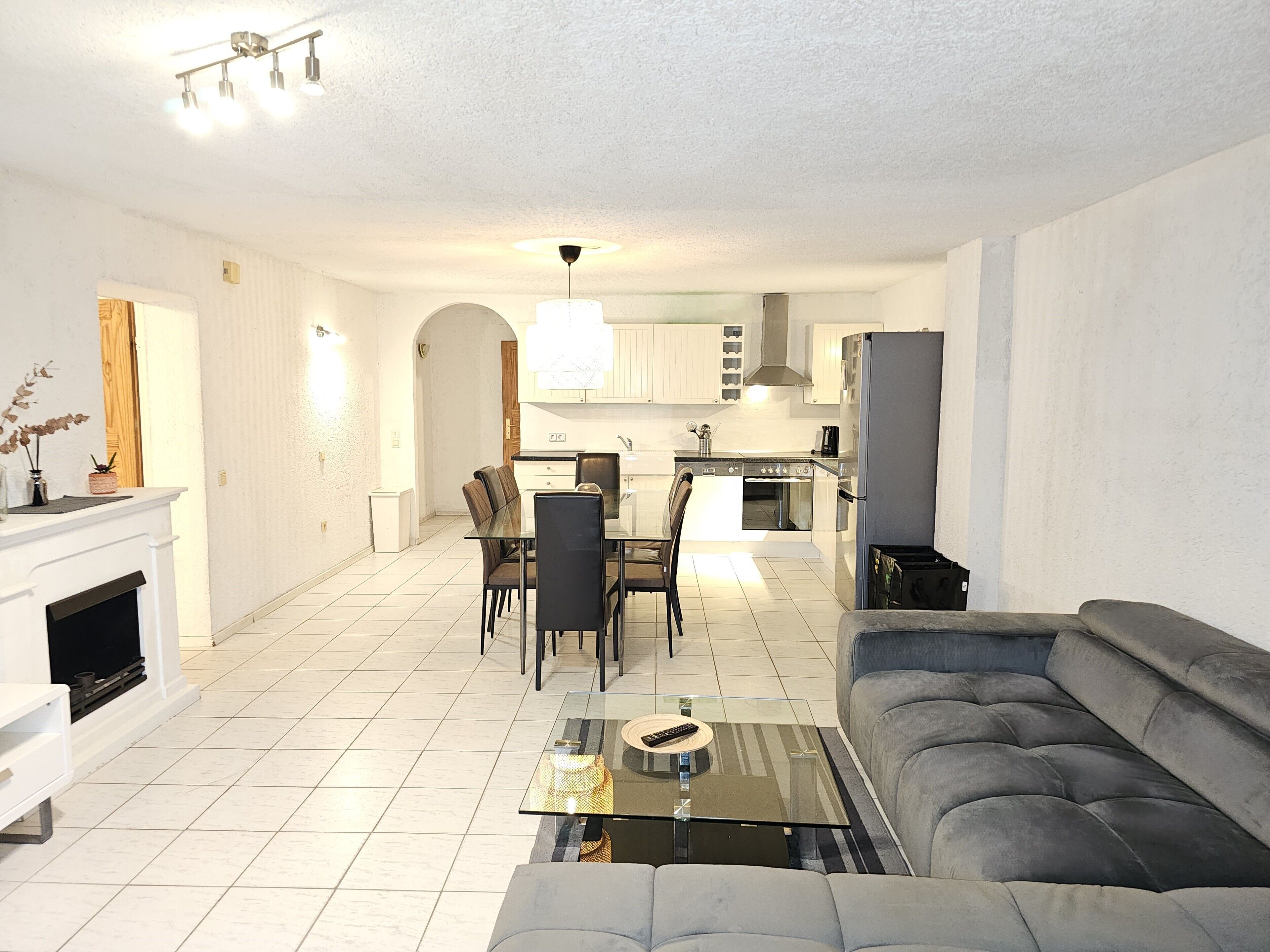 A spacious living room with a grey sofa, a glass coffee table, and a dining area with chairs and a table.