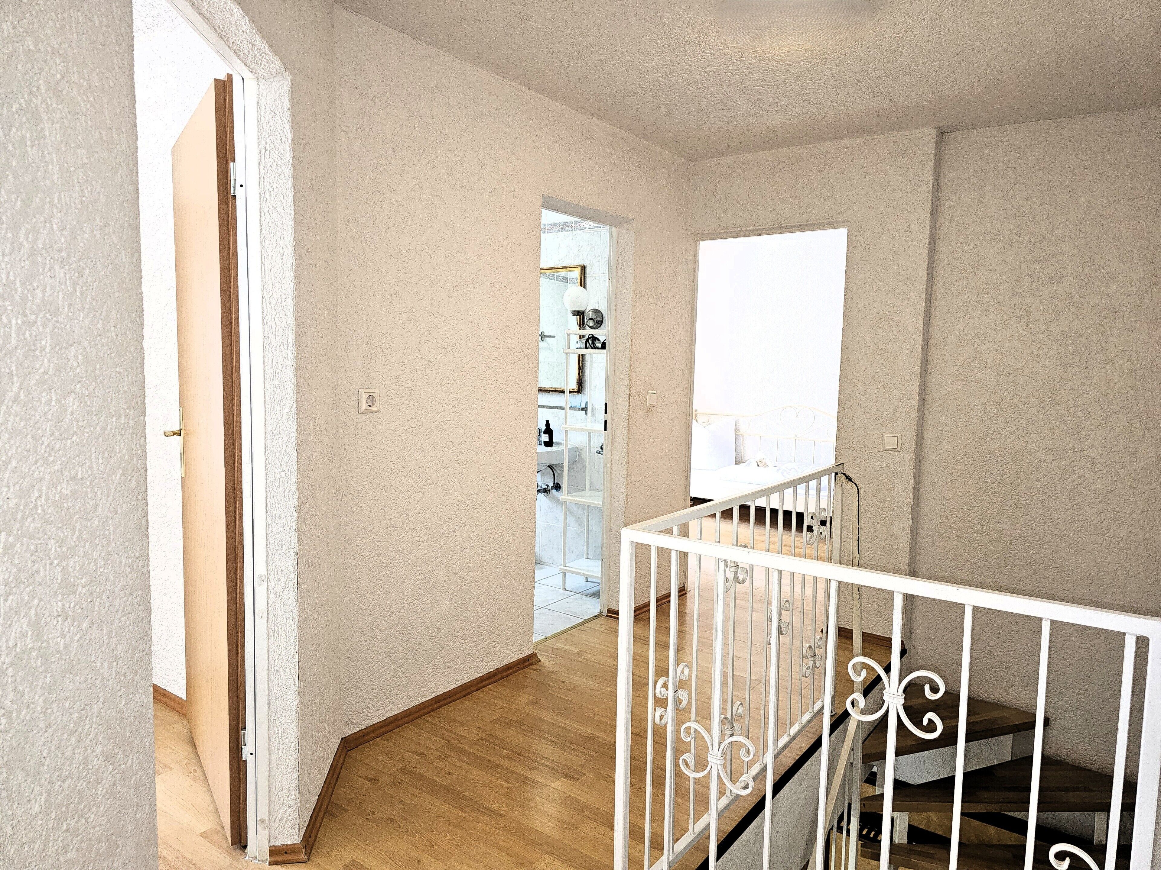 A hallway with a wooden floor, a white railing, and a door leading to a bathroom.