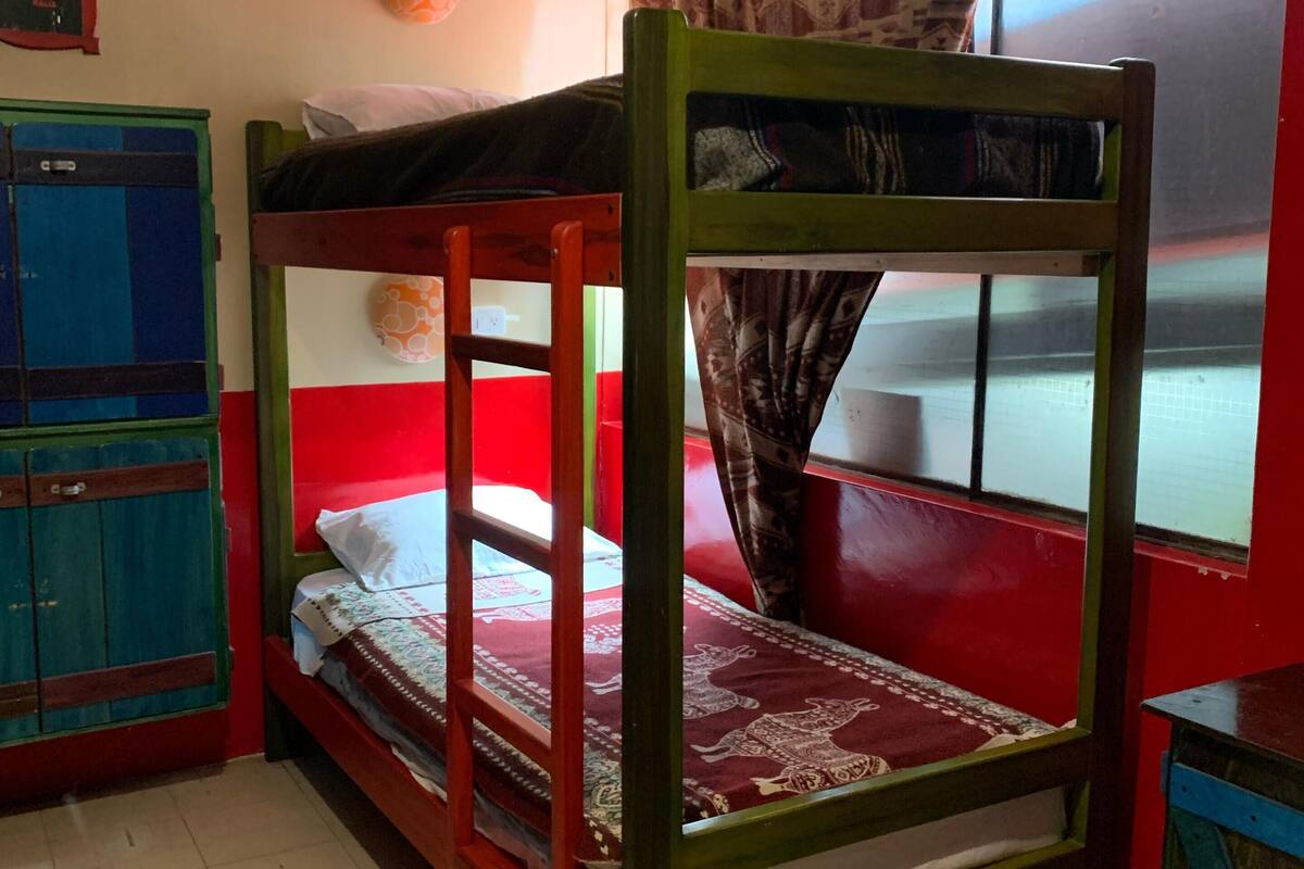 A bunk bed with a red and green frame, a small bedside table, and a window with patterned curtains.