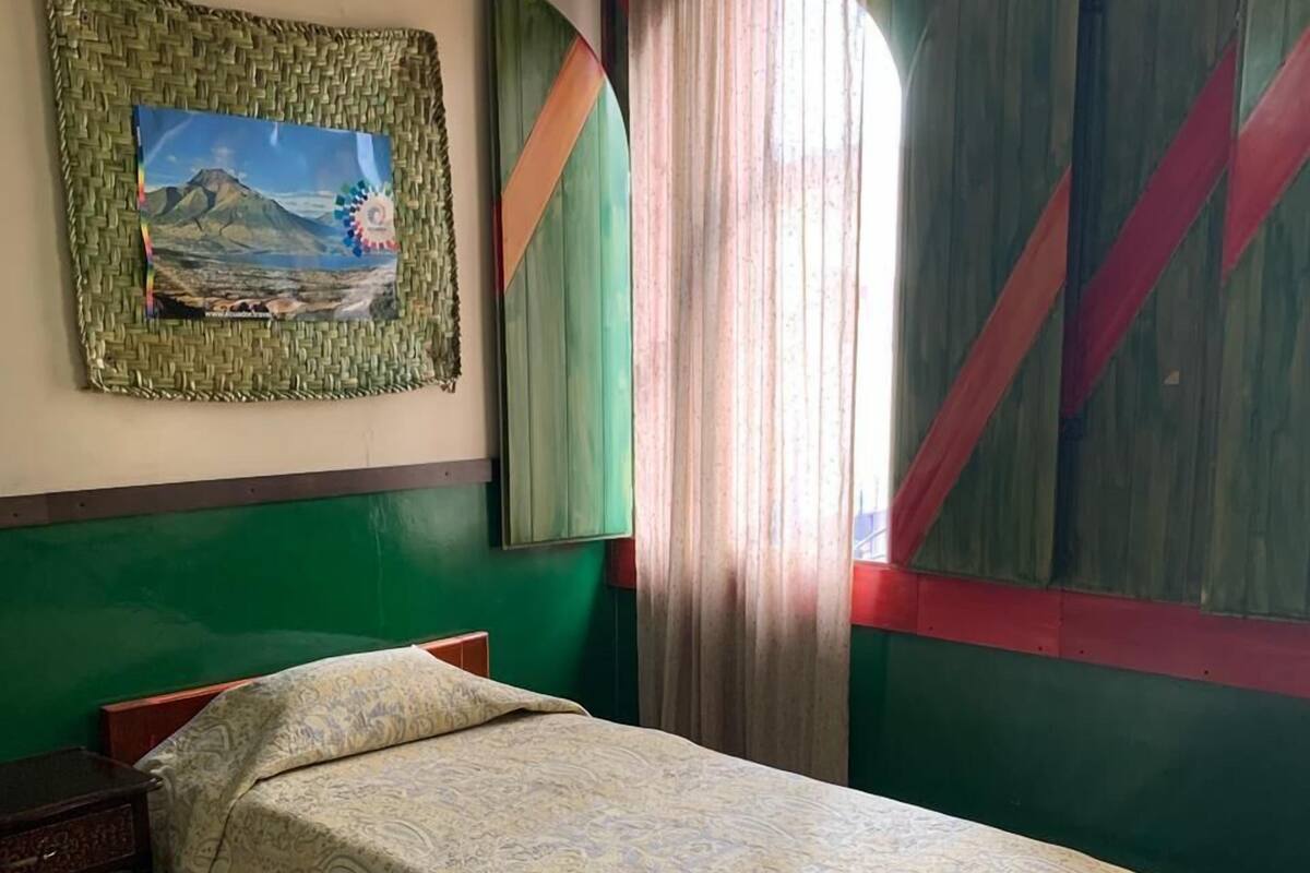 A single bed with a patterned bedspread in a room with green walls, a painting of a landscape, and a window with sheer curtains.