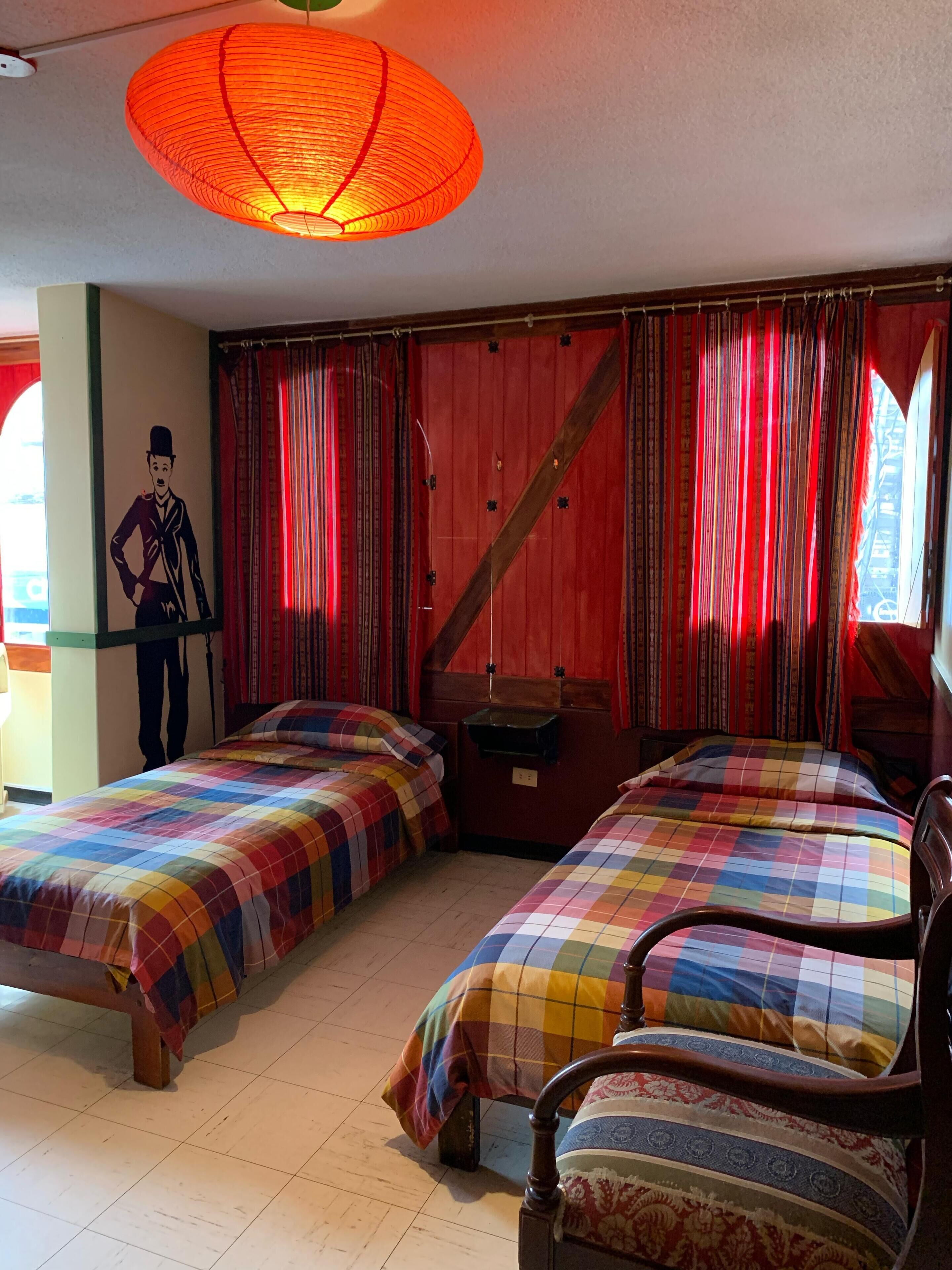 A room with two beds, a wooden chair, a hanging lamp, and a wall with a framed picture.