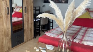 Une chambre avec une armoire en bois, un lit avec une literie rouge, un bureau avec une chaise, une bibliothèque et un tableau accroché au mur.