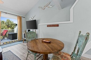 Une pièce avec une table ronde en bois, deux chaises, un téléviseur fixé au mur et un balcon offrant une vue sur les palmiers.