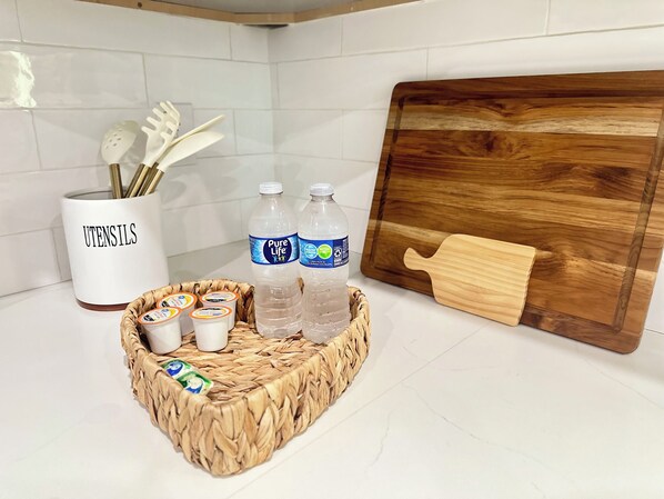 A kitchen counter with a white utensil holder, two water bottles, a wooden cutting board, and a woven basket containing yogurt containers.