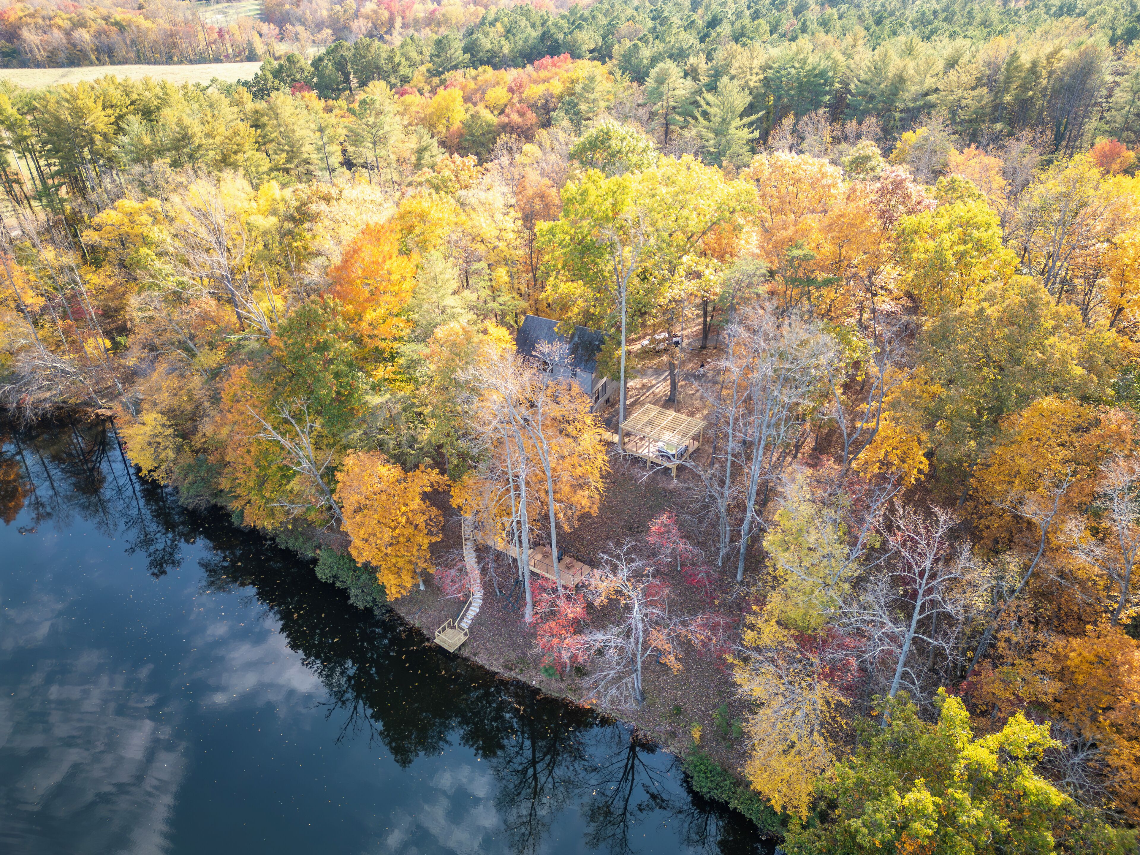 Luftaufnahme eines bewaldeten Gebiets mit einem Fluss, Bäumen in Herbstfarben und einer kleinen Gebäudesstruktur.