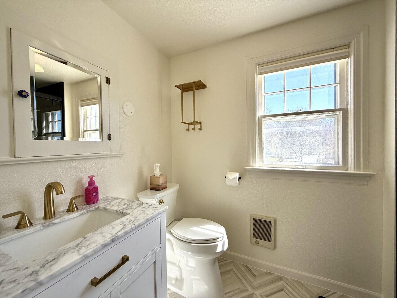 Une salle de bain avec des toilettes blanches, un lavabo avec des robinets dorés, un miroir et une fenêtre donnant sur des arbres.