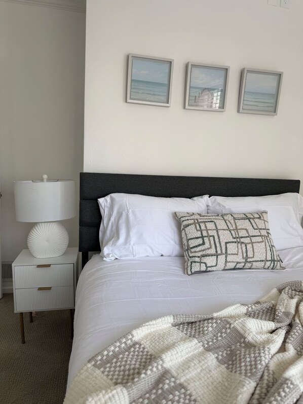 Una cama bien hecha con sábanas blancas, un cabecero oscuro y una manta decorativa estampada. Hay una mesita de noche con una lámpara y un adorno. Tres cuadros enmarcados están colgados en la pared sobre la cama.