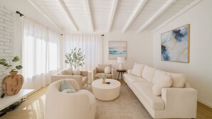 A modern living room with a white sofa, a round coffee table, and a white armchair. There is a large window with sheer curtains, a plant, and a painting on the wall. - Coastal Charm Awaits: Book Your Getaway Today! (Manhattan Beach)
