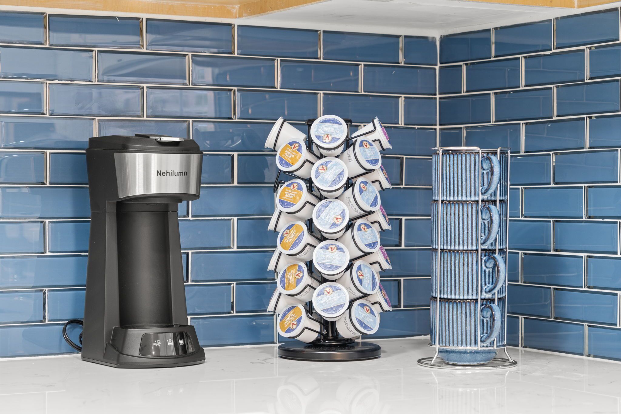 Una cafetera, un dispensador con varios pods de café y un recipiente alto frente a una pared de azulejos azules.