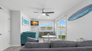 A modern living room with a grey sofa, teal accent chair, and a ceiling fan. A large window offers a view of the outdoors. - Cozy Condo in Siesta key with boat dock (Sarasota)