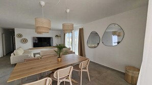 Een moderne woonkamer met een houten eettafel, rieten stoelen, een bank en een televisie. Er zijn twee wandspiegels en een plafondlamp.