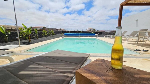A pool area with lounge chairs and a bottle of drink on a wooden table. - Villa in Lajares with a heated pool, garden, patio, and barbecue (La Oliva)