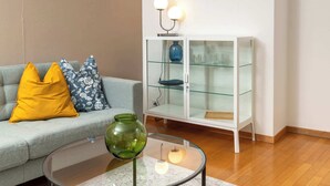 Een moderne woonkamer met een grijze bank, kussens met een geel en blauw patroon, een glazen salontafel met een groene vaas, en een witte kast met glazen planken en wandlampen.