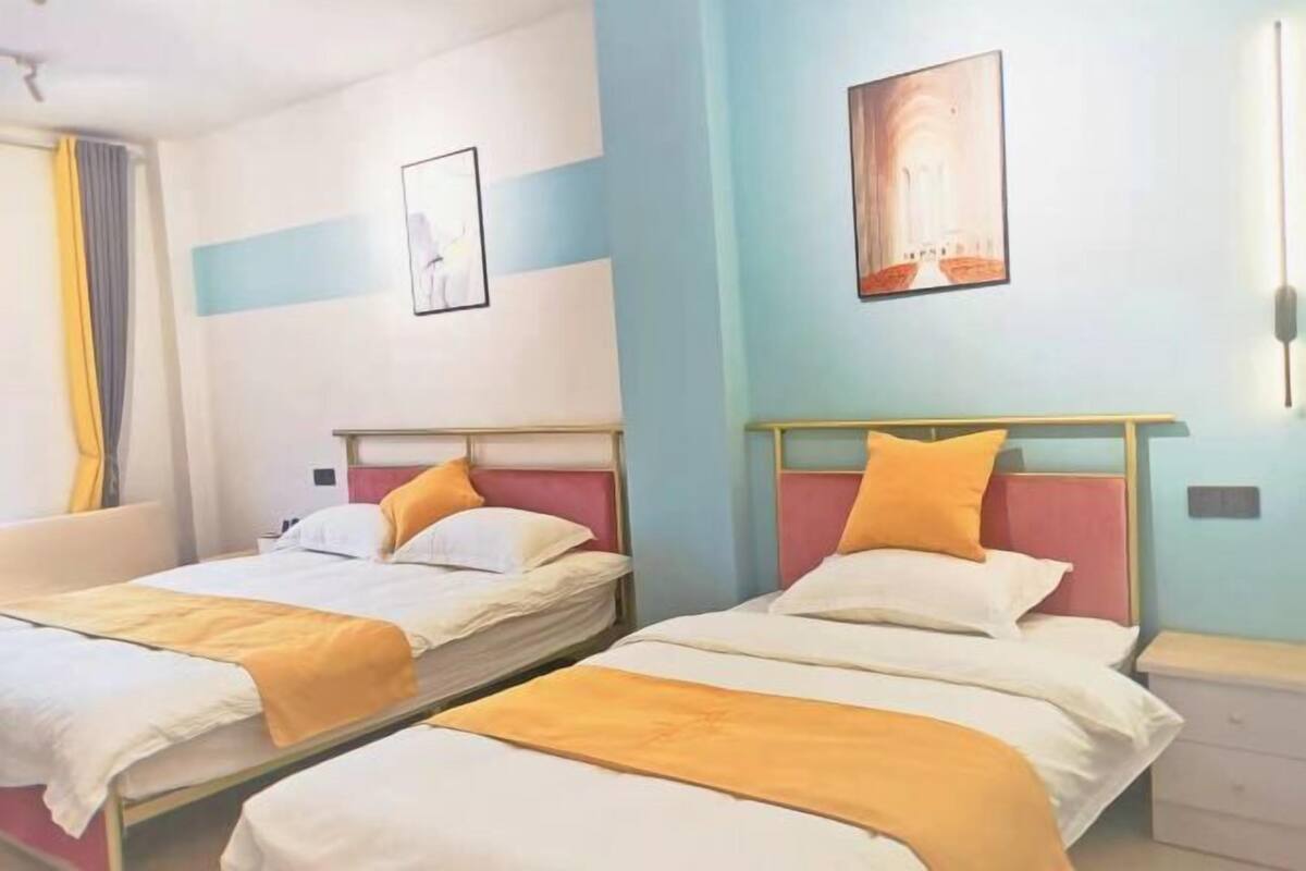 Two single beds with white linens and orange accents, a wall-mounted light, and a framed picture.
