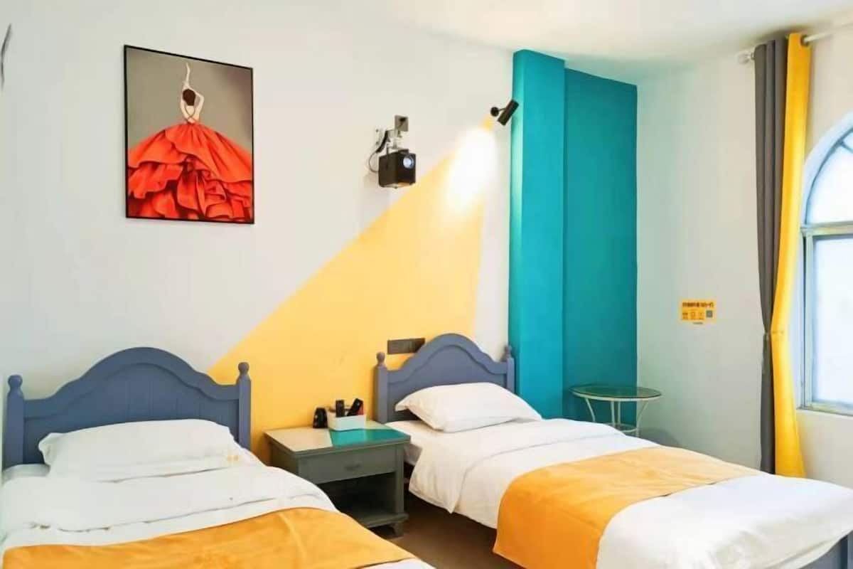 A hotel room with two beds, a nightstand, a small table, a painting, and a window with curtains.