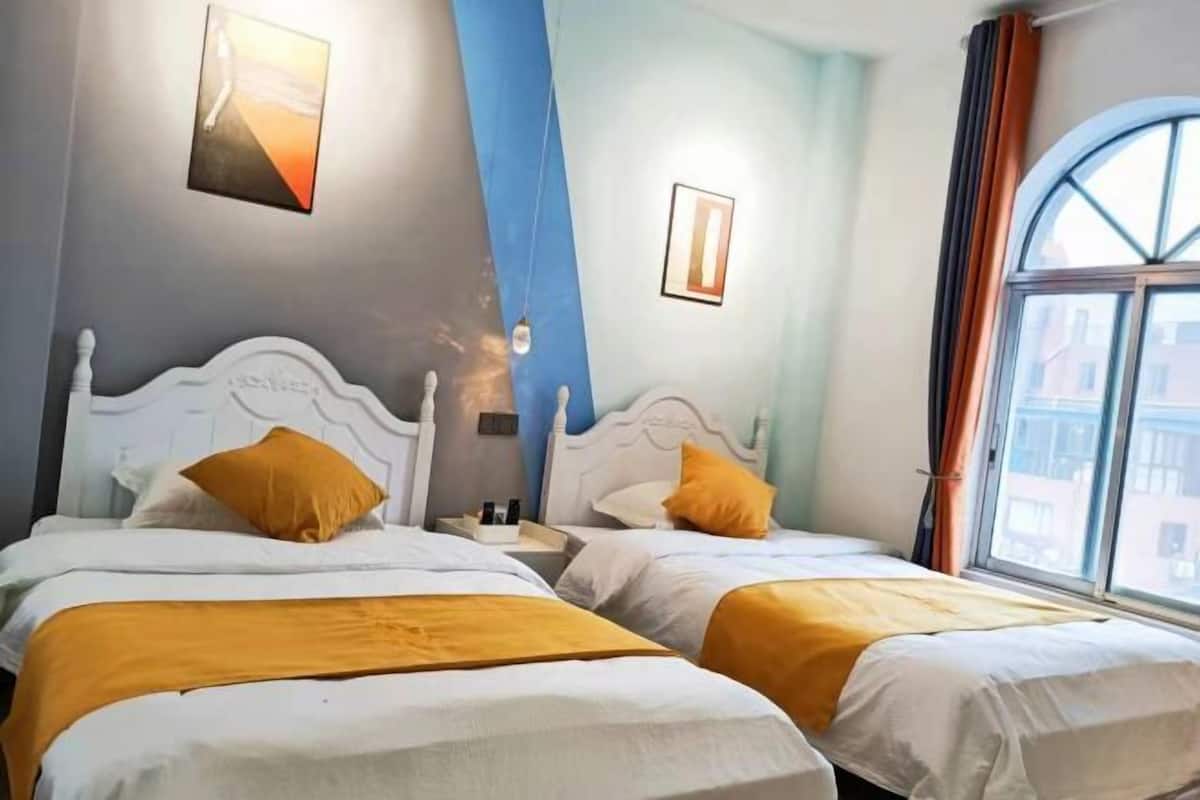 A hotel room with two beds, each with white headboards and orange bedding. There is a window with curtains, a framed picture on the wall, and a blue accent wall.