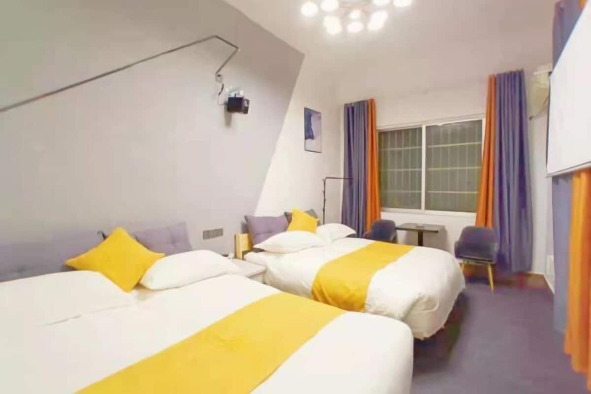 A hotel room with two beds, a desk, a chair, a window with curtains, and a ceiling light fixture.
