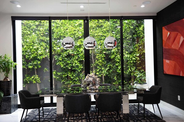 A modern dining area with a glass table, black chairs, and a view of lush greenery through large windows. - Melrose Mansion - Pool, Rooftop + Firepit & Grill (West Hollywood)