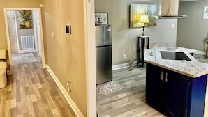 Una cocina moderna con refrigerador empotrado, estufa y pileta. Hay una pequeña isla con cubierta de mármol y una lámpara. El piso es de madera y se ve parte de la sala.