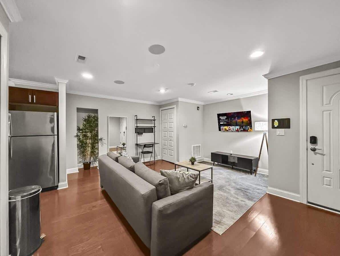 A modern living room with a grey sofa, a dining area, a kitchen with a stainless steel refrigerator, and a large TV on the wall.