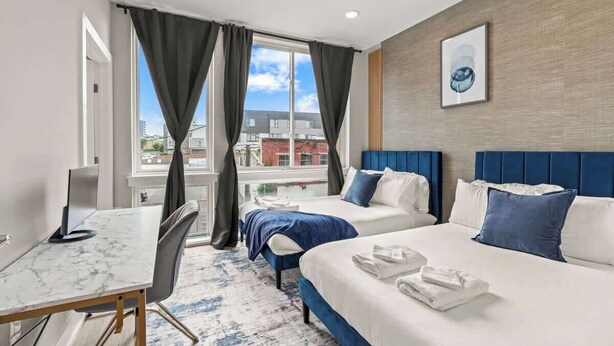 Una habitación de hotel con dos camas, un escritorio y un amplio ventanal con cortinas.