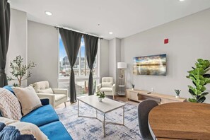 Een moderne woonkamer met een blauwe bank, een witte salontafel, een grijze fauteuil en een houten bijzettafeltje. Er hangt een schilderij aan de muur en er is een raam met gordijnen.
