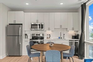 A modern kitchen with white cabinets, stainless steel appliances, and a round wooden dining table with four chairs. - Luxury 1BD | 3 Beds | Northern Liberties (Philadelphia)