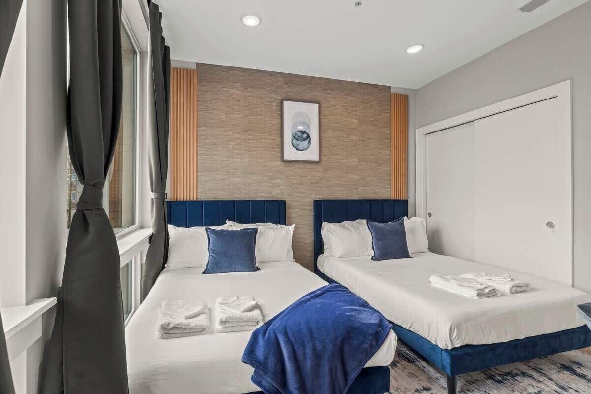Ein Hotelzimmer mit zwei Betten, einem großen Fenster mit Vorhängen, einem gerahmten Bild an der Wand und einer Tür.