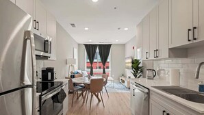 A modern kitchen with stainless steel appliances, a dining area with a wooden table and chairs, and a view of a red building through large windows. - Luxury 1BD | 2 Beds | Northern Liberties (Philadelphia)
